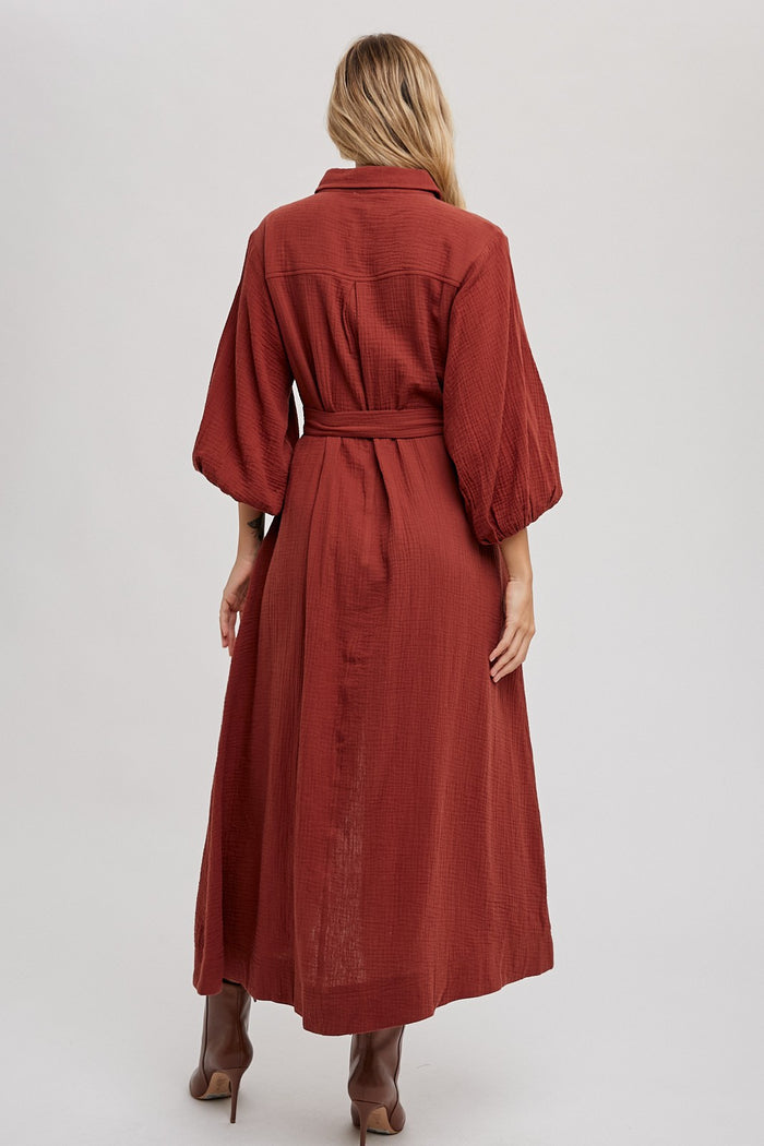 woman wearing a long rust-colored button-up dress with puff sleeves and a tied waist, shown from the back