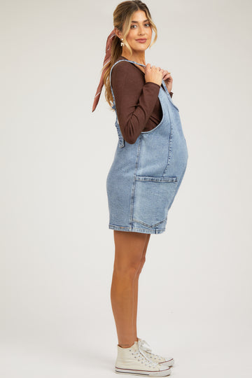 woman wearing a light blue denim maternity overall dress and a brown long sleeve top, showcasing her baby bump