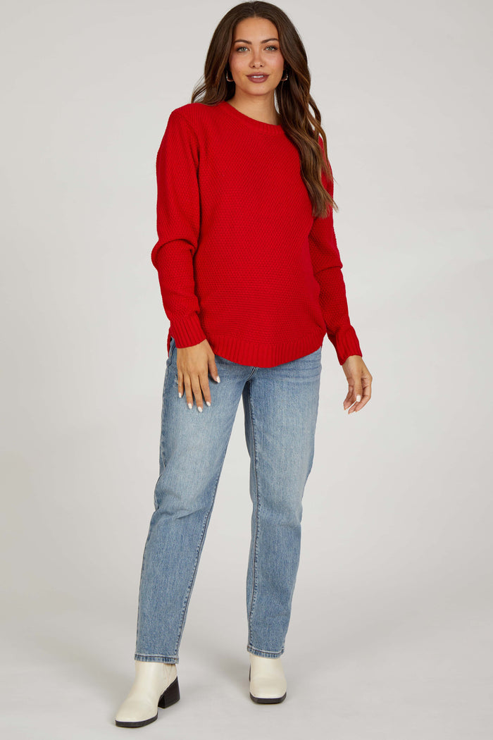 woman wearing a red knitted maternity sweater with side slits, paired with light wash jeans and ankle boots