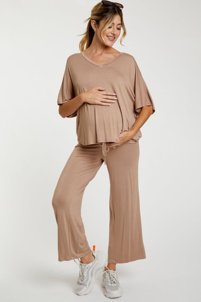 woman wearing a beige maternity top and matching pants, holding her baby bump and smiling