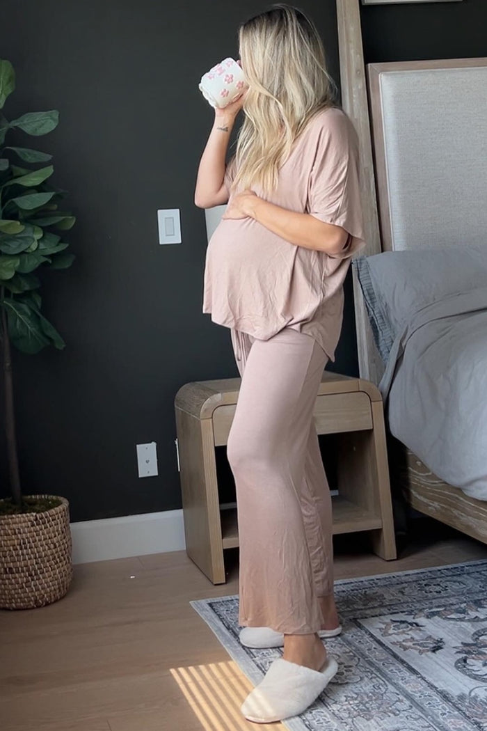 woman wearing a blush pink maternity loungewear set with a visible baby bump, holding a coffee cup and wearing slippers