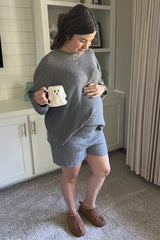 woman wearing a grey maternity loungewear set with a loose top and shorts, holding a coffee mug and showing her baby bump
