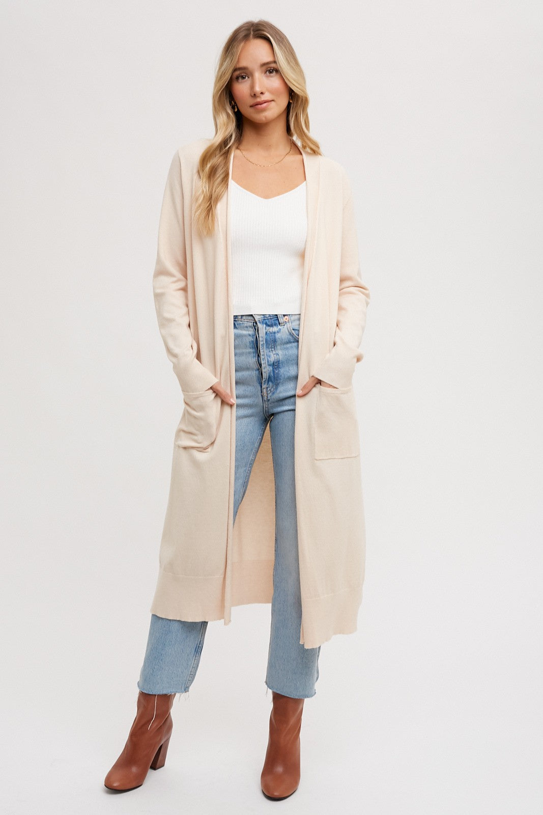 woman wearing a long beige cardigan over a white v-neck top and blue high-waisted jeans, standing with hands in pockets