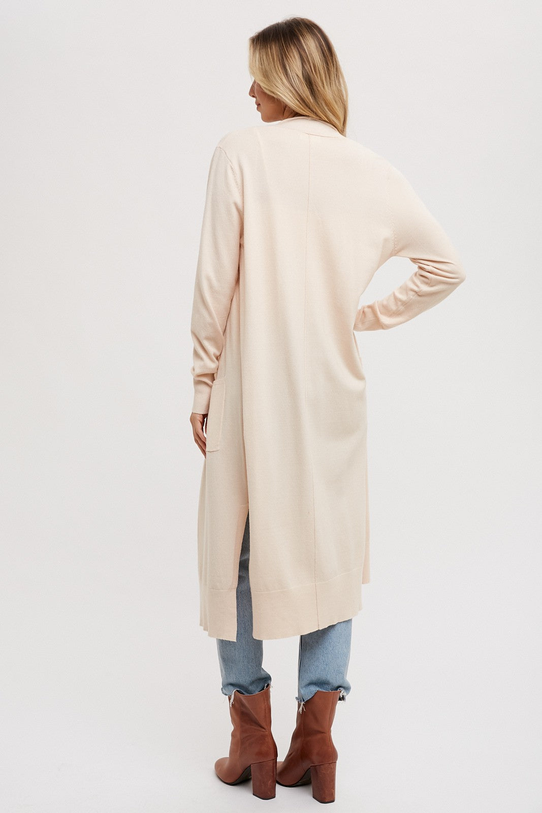 woman wearing a light pink long cardigan with side slits, standing with her back towards the camera, paired with jeans and ankle boots