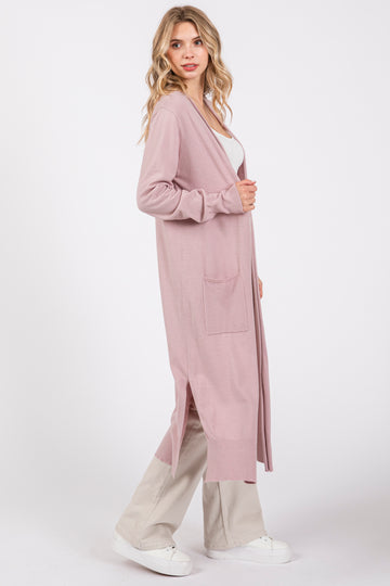 woman wearing a long pink cardigan with side slits and pockets, styled with a white tank top and light beige pants