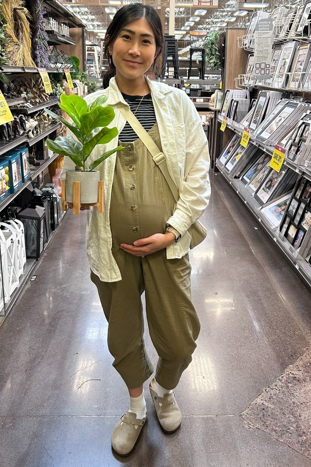 woman wearing a light green maternity jumpsuit and holding a potted plant while showing her baby bump in a store