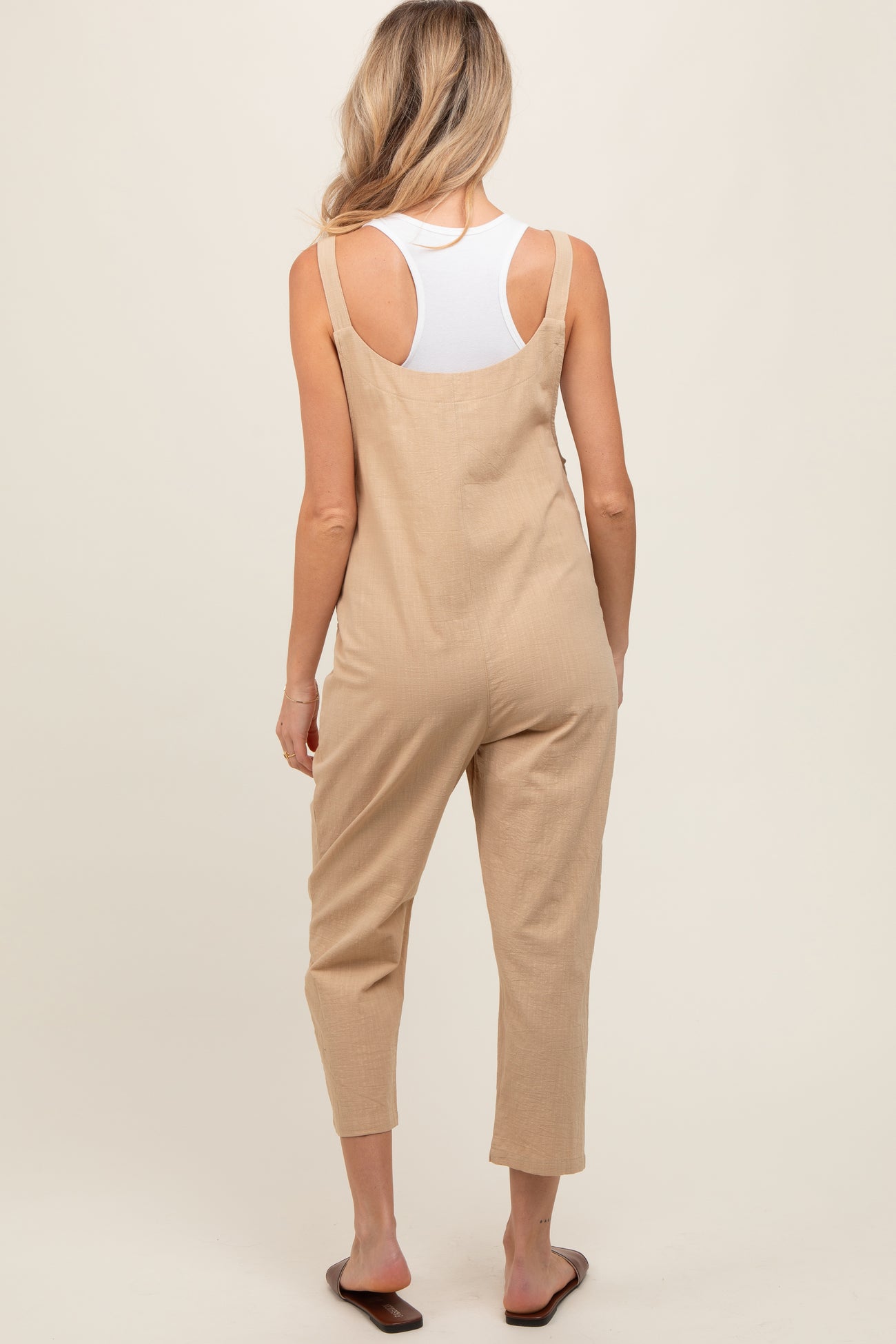 woman wearing a beige maternity jumpsuit with a relaxed fit and adjustable straps, seen from the back
