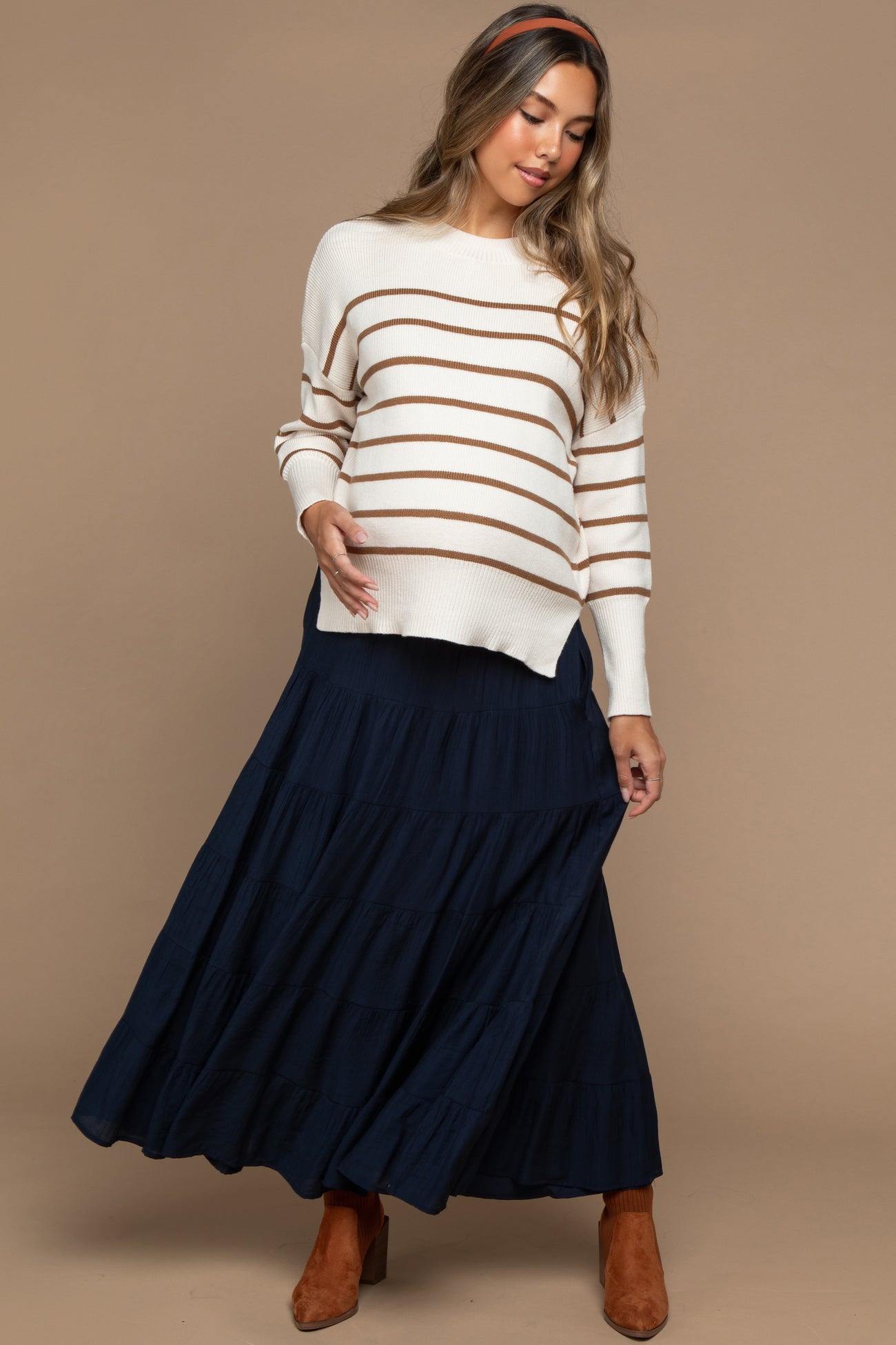 woman wearing a cream and brown striped maternity sweater and a navy tiered maxi skirt, holding her baby bump