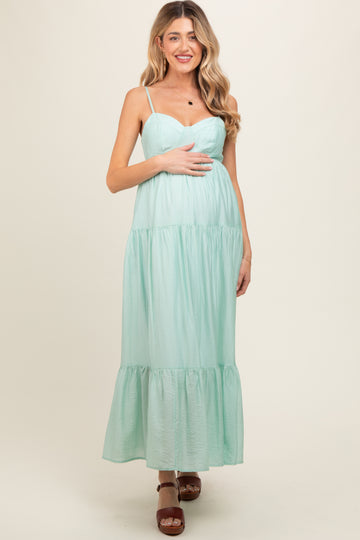 woman wearing a mint green maternity maxi dress with spaghetti straps and tiered detailing, holding her baby bump