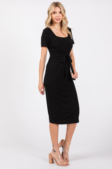 woman wearing a black short sleeve tie-waist dress, standing against a plain background