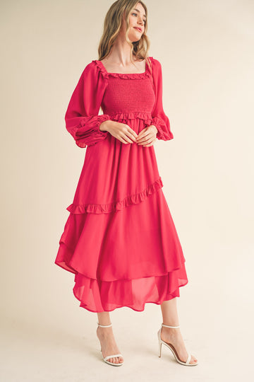 woman wearing a bright pink long sleeve dress with ruffles and a tiered hem, standing and smiling
