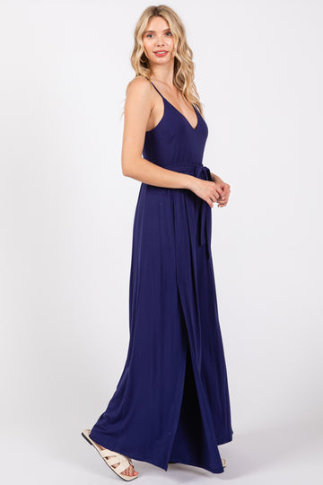 woman wearing a navy blue maxi dress with a deep v-neckline and a tie belt, standing in profile with a slit at the side