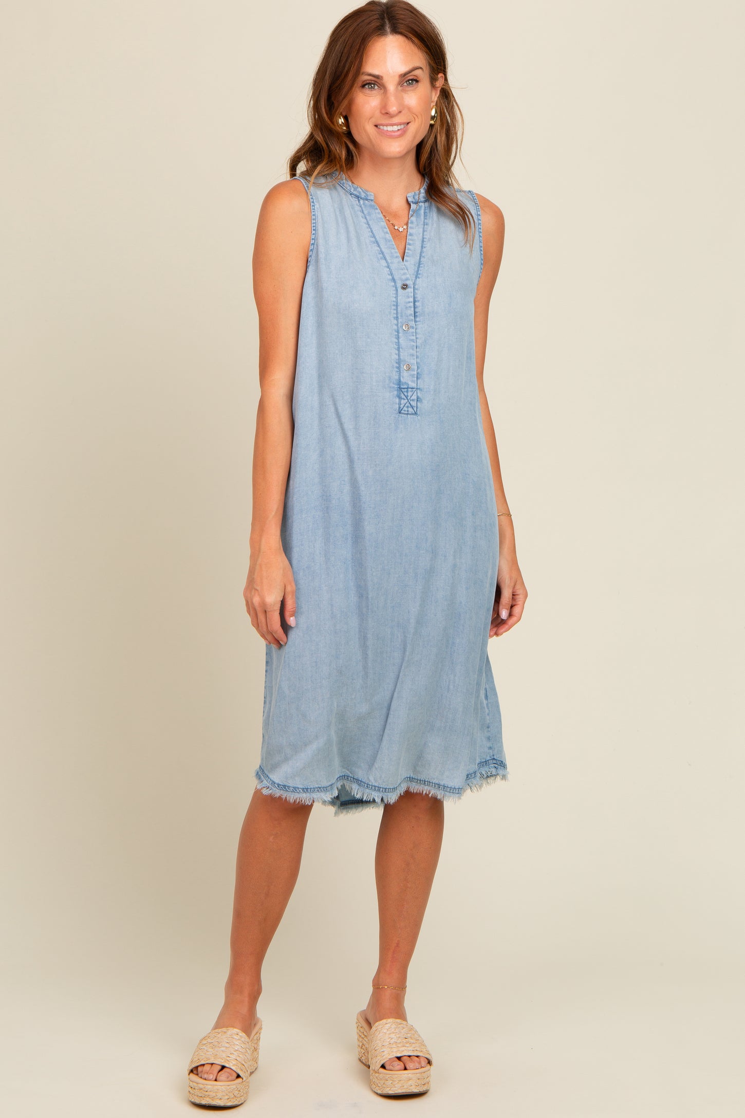 women wearing a light blue sleeveless denim dress with a button-up front and frayed hem, standing in a neutral background