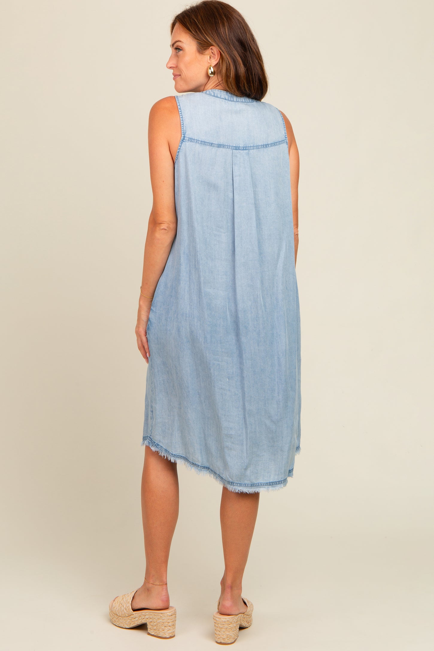 woman wearing a light wash sleeveless denim dress with a back pleat and frayed hem, styled with espadrille sandals