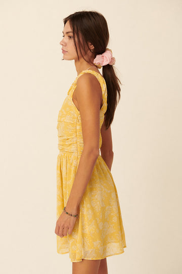 woman wearing a yellow sleeveless dress with a floral pattern and gathered details, standing sideways with hair in a ponytail